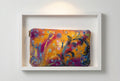 A rectangular zippered pouch or small bag with an abstract painterly print in orange, yellow, purple, blue, and black, photographed on a white tabletop with part of a gray chair and a small orange item visible at the right edge.