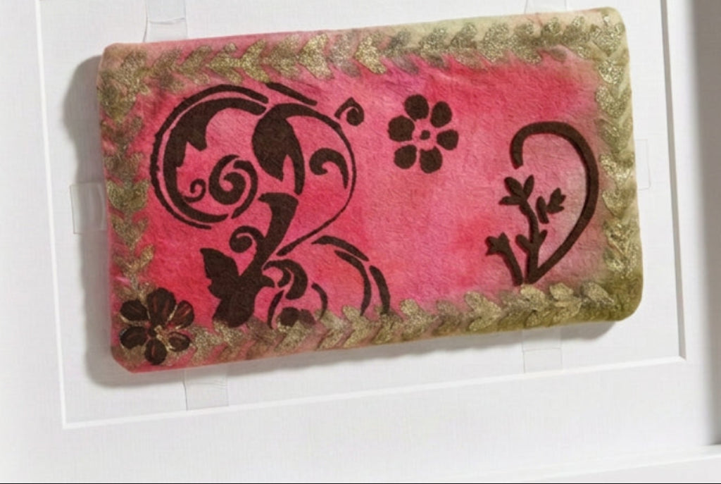 A rectangular fabric pouch or clutch with a pink center, black floral and scroll motifs, and a distressed olive-brown border, photographed on a white surface.