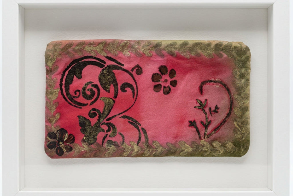 A rectangular fabric pouch or clutch with a pink center, black floral and scroll motifs, and a distressed olive-brown border, photographed on a white surface.