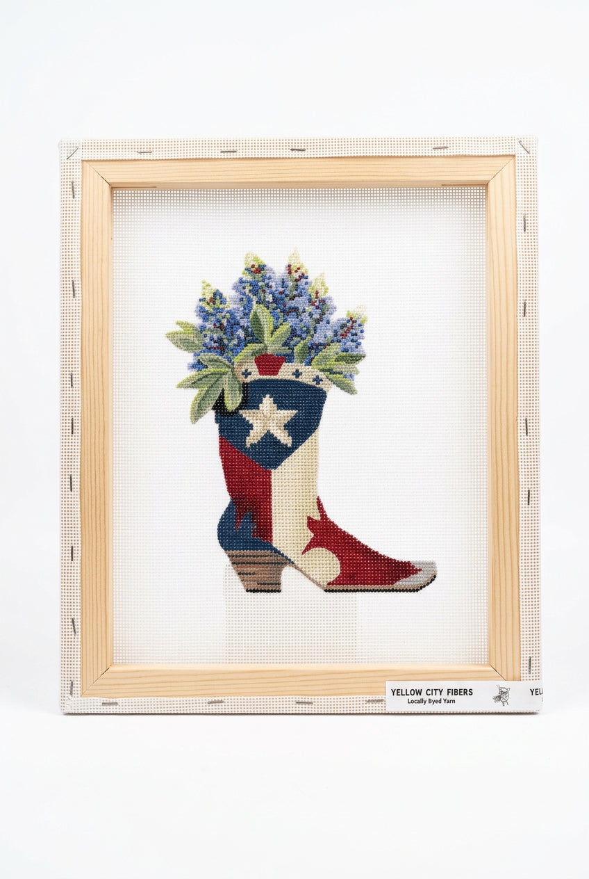 A needlepoint canvas featuring a cowboy boot filled with purple flowers, printed on white A4 mesh. The canvas is branded with 'Yellow City Fibers' and has a clip at the top, photographed on a white surface.