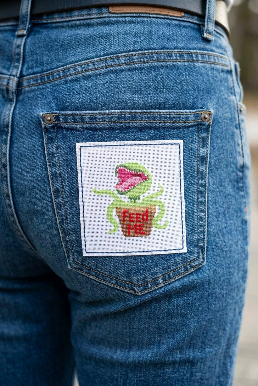 A square white needlepoint canvas featuring a pixelated green plant monster with its mouth wide open, sitting in a brown pot with red text that reads 'Feed Me.' The canvas is photographed against a light beige background, with the number '25209' printed in black near the bottom left.
