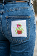 A square white needlepoint canvas featuring a pixelated green plant monster with its mouth wide open, sitting in a brown pot with red text that reads 'Feed Me.' The canvas is photographed against a light beige background, with the number '25209' printed in black near the bottom left.
