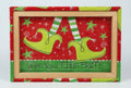 A festive needlepoint canvas featuring a red background with green stars, striped green-and-white socks, two yellow-green elf shoes, and the text 'Wipe Your Elfin FEET !!' on a green banner at the bottom.