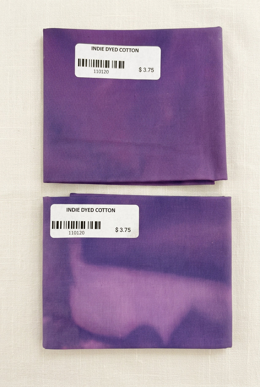 Two folded square pieces of purple indigo-dyed cotton fabric with white price labels, displayed on a white surface.