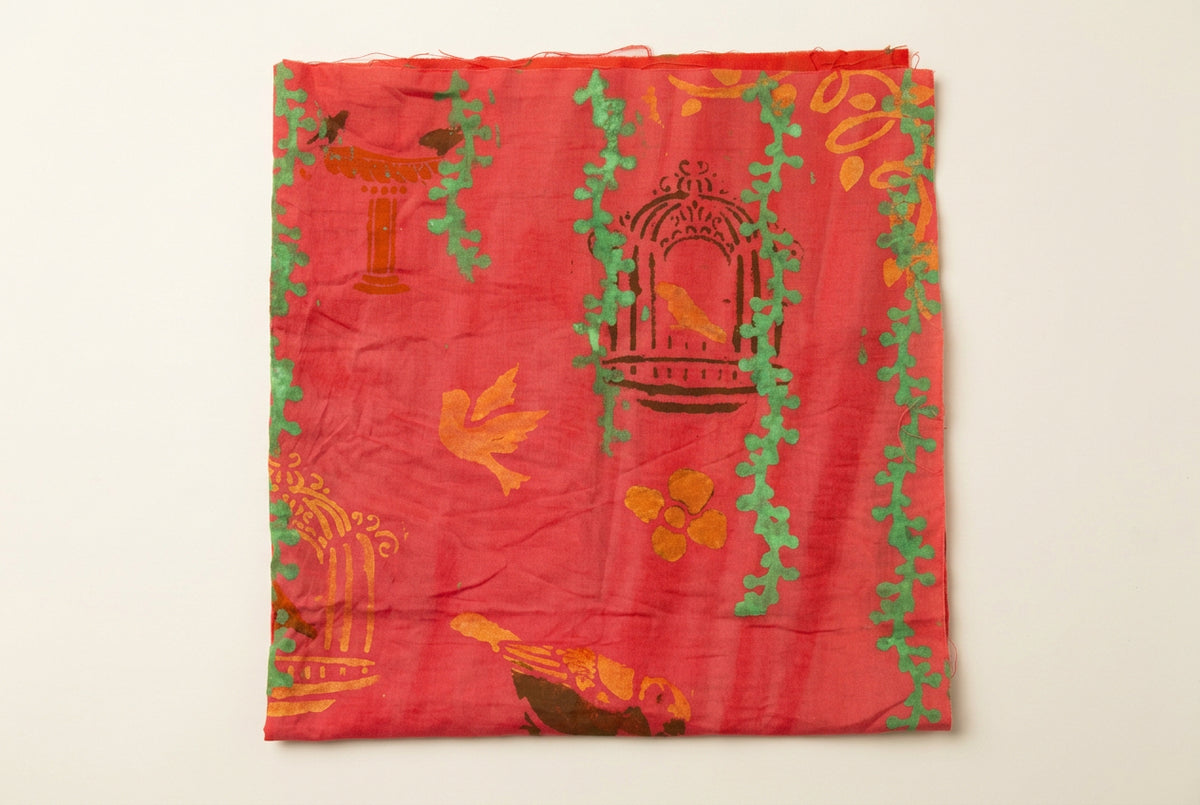 A square piece of coral pink fabric with a printed pattern of gold birds, green ivy vines, and brown birdcages, laid flat on a white desk.