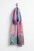 A rectangular scarf laid flat on a dark desk, featuring a pink and purple tie-dye pattern with teal, orange, and black floral motifs concentrated along the bottom edge.
